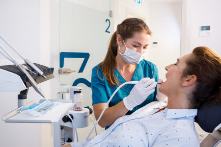 Eine Zahnmedizinische Fachangestellte behandelt eine Patientin im Behandlungsstuhl. Beide befinden sich in einem modernen Behandlungszimmer, die Fachangestellte trägt Maske und Handschuhe.