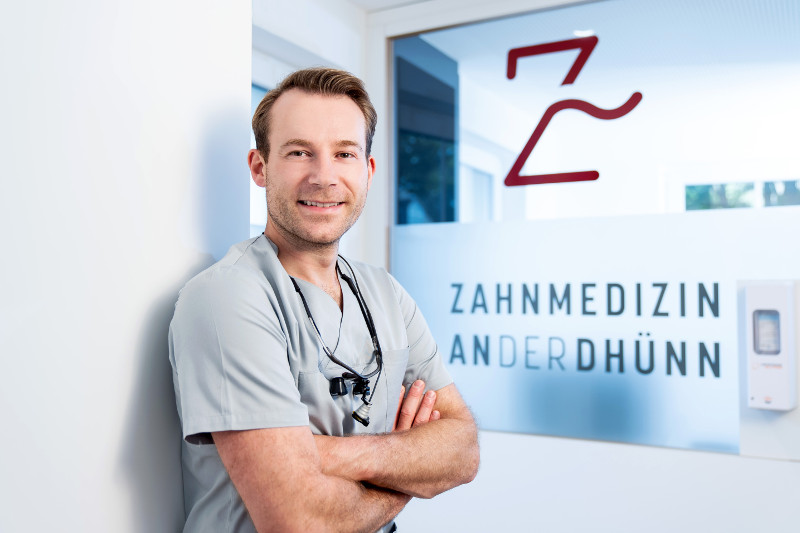 Dr. Benjamin Schulte, Zahnarzt der Praxis „Zahnmedizin an der Dhünn“, lehnt lächelnd mit verschränkten Armen an einer Wand. Im Hintergrund ist das Logo der Praxis auf einer Glastür zu sehen. Die Atmosphäre wirkt professionell und freundlich.