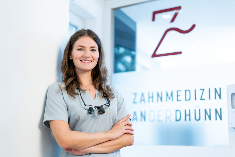 Zahnärztin Thalia Hampl von der Praxis „Zahnmedizin an der Dhünn“ lehnt lächelnd mit verschränkten Armen an einer Wand. Im Hintergrund ist das Logo der Praxis auf einer Glastür zu sehen. Die Atmosphäre wirkt professionell und freundlich.