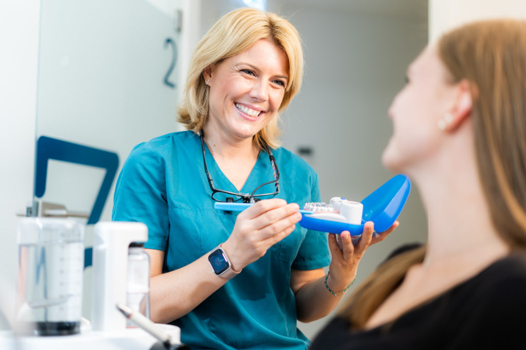 Eine zahnmedizinische Assistentin in blauer Kleidung erklärt einer Patientin mit einem Zahnmodell in einer blauen Box etwas, während sie lächelt.
