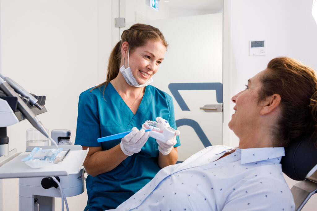 Eine zahnmedizinische Assistentin in blauer Kleidung erklärt einer Patientin mit einem Zahnmodell und einer medizinischen Zahnbürste etwas, während sie lächelt.