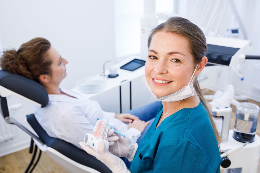 Professioneller Mundgesundheitscheck – ZFA erklärt Patientin die richtige Zahnpflege mit einem Modell. Zahnmedizinische Fachangestellte (ZFA) in blauer Kleidung lächelt in die Kamera, bevor sie einer Patientin auf dem Behandlungsstuhl ein Zahnmodell zeigt, welches sie in der Hand hält. Im Hintergrund ist eine moderne Zahnarztpraxis zu sehen.