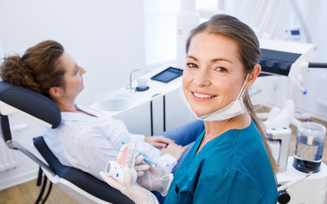 Zahnmedizinische Fachangestellte (ZFA) in blauer Kleidung lächelt in die Kamera, bevor sie einer Patientin auf dem Behandlungsstuhl ein Zahnmodell zeigt, welches sie in der Hand hält. Im Hintergrund ist eine moderne Zahnarztpraxis zu sehen.