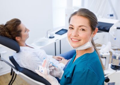 Zahnmedizinische Fachangestellte (ZFA) in blauer Kleidung lächelt in die Kamera, bevor sie einer Patientin auf dem Behandlungsstuhl ein Zahnmodell zeigt, welches sie in der Hand hält. Im Hintergrund ist eine moderne Zahnarztpraxis zu sehen.