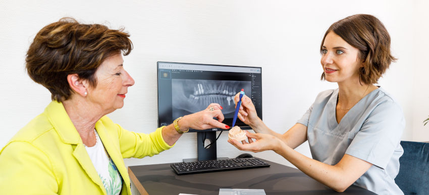Eine Zahnärztin in hellgrauer Kleidung, Dr. Sabine Schulte, erklärt einer älteren Patientin anschaulich ein Zahnmodell. Beide sitzen an einem Schreibtisch, im Hintergrund ist auf einem Monitor ein Röntgenbild eines Gebisses zu sehen. Die Zahnärztin zeigt mit einem Stift auf das Modell, während die Patientin aufmerksam zuhört und das Modell berührt.