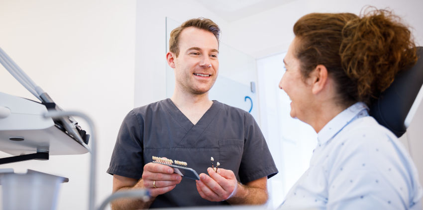 Dr. Schulte hält verschiedene Zahnersatz-Modelle in den Händen und berät eine Patientin, die auf einem Behandlungsstuhl sitzt. Beide wirken freundlich und entspannt.