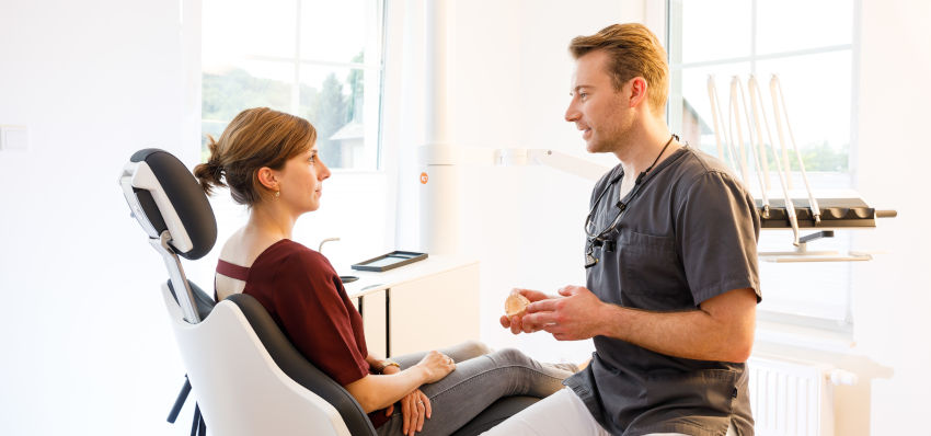 Dr. Benjamin Schulte, Zahnarzt in grauem Oberteil und weißer Hose, spricht in einem hellen Behandlungsraum mit einer Patientin, die entspannt in einem modernen Behandlungsstuhl sitzt. Er hält ein Zahnmodell in der Hand und erklärt anschaulich die Behandlungsmöglichkeiten. Im Hintergrund sind große Fenster, Geräte und helle Möbel zu sehen.