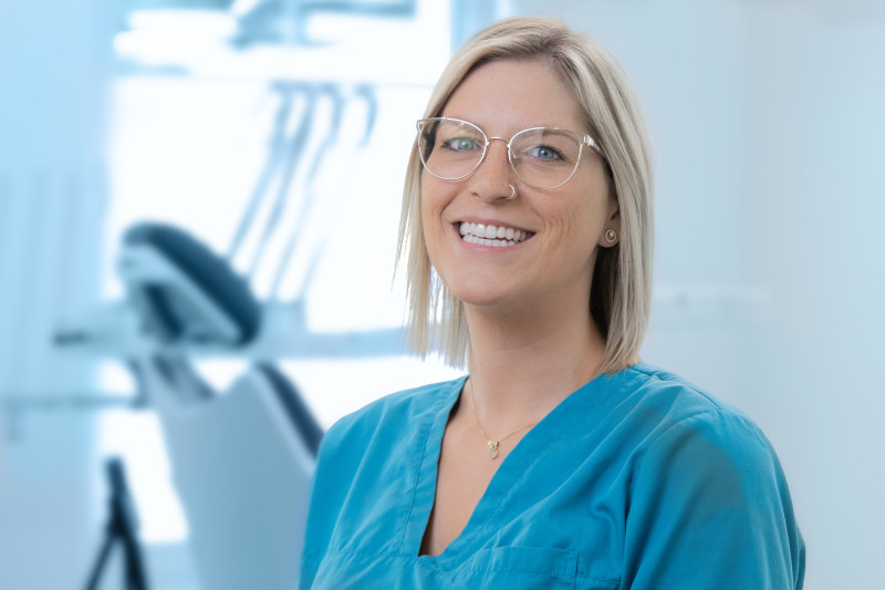 Johanna Ern steht lächelnd in einem hellen, modernen Behandlungszimmer einer Zahnarztpraxis. Sie trägt türkisfarbene Praxiskleidung und eine Brille mit transparentem Rahmen. Ihre blonden, glatten Haare sind kinnlang geschnitten. Sie trägt eine kleine Halskette und hat ein Nasenpiercing. Im Hintergrund ist unscharf ein zahnärztlicher Behandlungsstuhl mit Instrumenten zu erkennen. Die Szene wirkt freundlich und professionell.