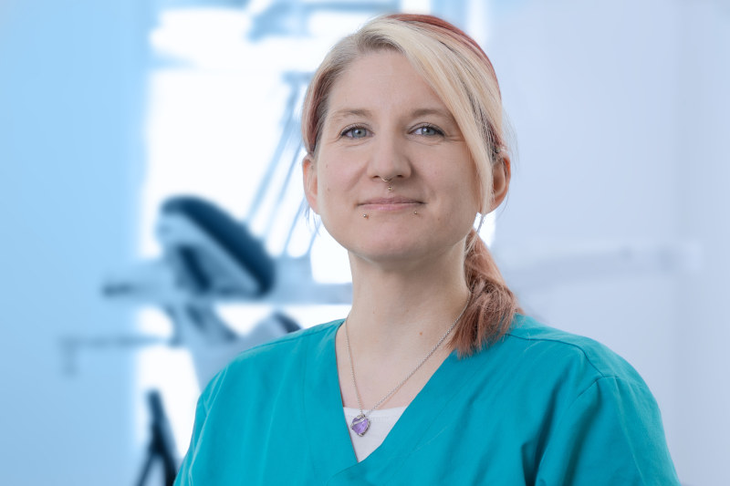 Nadine Mebus steht in türkisfarbener Praxiskleidung in einem modernen Behandlungszimmer einer Zahnarztpraxis. Sie hat rötlich-blonde Haare, die seitlich zum Zopf gebunden sind, und trägt mehrere Piercings im Gesicht sowie eine Halskette mit violettem Anhänger. Ihr Blick ist ruhig und freundlich. Im Hintergrund ist unscharf ein zahnärztlicher Behandlungsstuhl mit Instrumenten zu erkennen. Die Atmosphäre wirkt professionell und angenehm.