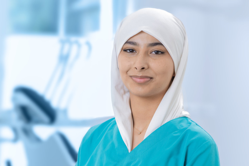 Die Auszubildende Rukhsar Qaderi steht vor einem hellen, modern ausgestatteten Behandlungszimmer in einer Zahnarztpraxis. Sie trägt türkisfarbene Praxiskleidung und ein weißes Kopftuch, das ordentlich um ihren Kopf geschlungen ist. Sie blickt ruhig und freundlich in die Kamera. Im Hintergrund sind unscharf ein Behandlungsstuhl und medizinisches Gerät zu erkennen. Die Atmosphäre wirkt professionell und einladend.